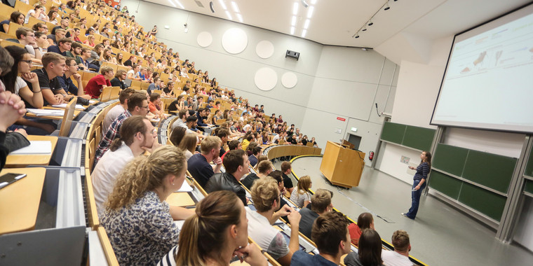 Vorlesung im Hörsaal Viele Studierende sitzen bei einer Vorlseung im Hörsaal. Vorne steht eine Dozentin.