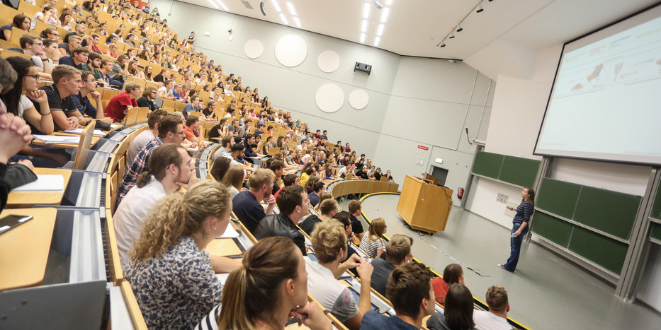 Vorlesung im Hörsaal Viele Studierende sitzen bei einer Vorlseung im Hörsaal. Vorne steht eine Dozentin.