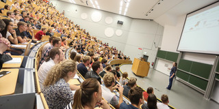Vorlesung im Hörsaal Viele Studierende sitzen bei einer Vorlseung im Hörsaal. Vorne steht eine Dozentin.
