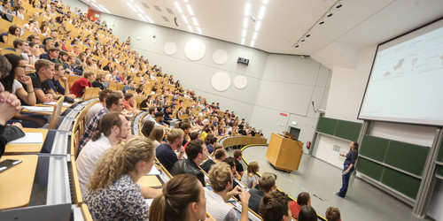 Vorlesung im Hörsaal Viele Studierende sitzen bei einer Vorlseung im Hörsaal. Vorne steht eine Dozentin.