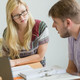 MatheHelpdesk Nahaufnahme von zwei Personen, die an einem Tisch sitzen. Die Person rechts hat Unterlagen vor sich liegen und spricht. Die Person links hört der anderen Person konzentriert zu.
