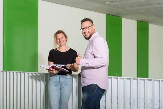 Studierende vor Geländer, Daumen hoch Zwei Studierende stehen vor einem Geländer, einer streckt den Daumen hoch.