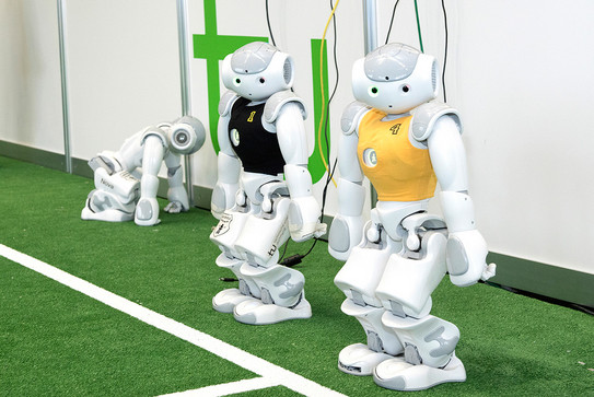 Three small white robots, one in a black jersey, one in a yellow jersey, standing next to a indoor soccer field with the green TU logo in the background.
