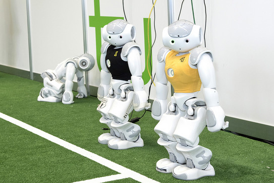 Three small white robots, one in a black jersey, one in a yellow jersey, standing next to a indoor soccer field with the green TU logo in the background.
