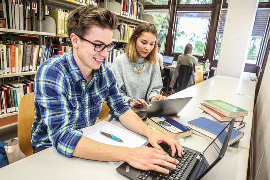Zwei Studierende sitzen in der Bibliothek und lernen am Laptop. 