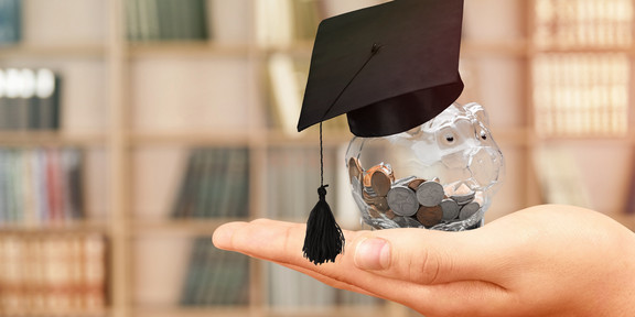 A transparent piggy bank full of coins and with a doctoral cap is held in front of a book shelf. 
