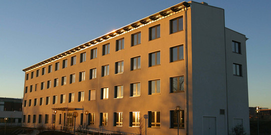 Gebäude E-Technik: Ein hohes weißes Gebäude, das von der Sonne angestrahlt wird. Vor dem Gebäude eine Rampe und über dem Eingang ein Vordach.