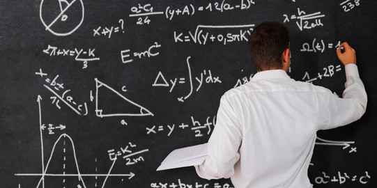 A man is seen from behind. He is writing formulas on a chalkboard