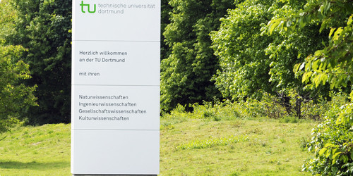 Ein weißes Begrüßungsschild der TU Dortmund steht auf einem grünen Rasen.