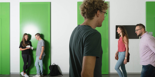 Studierende im Gehen Zwei Studierende lehnen an der Wand, zwei kommen von rechts ins Bild und einer geht nach rechts.
